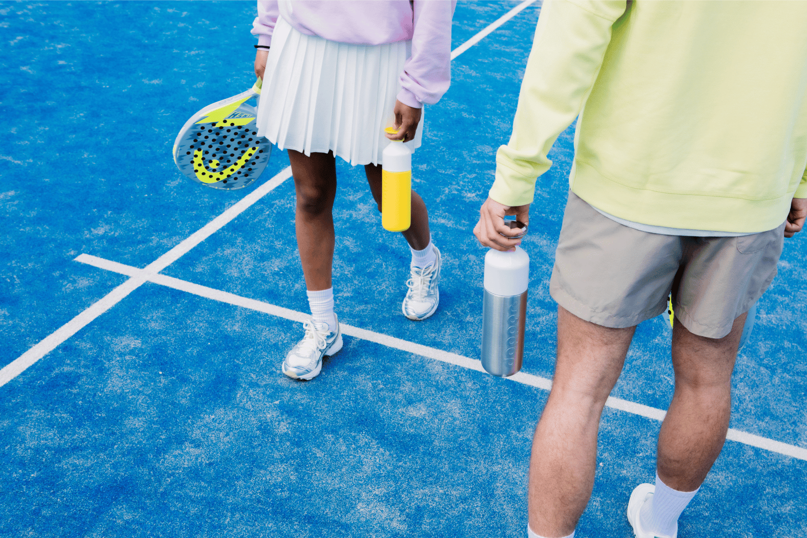 Two people holding padel rackets and Dopper bottles on a blue padel court