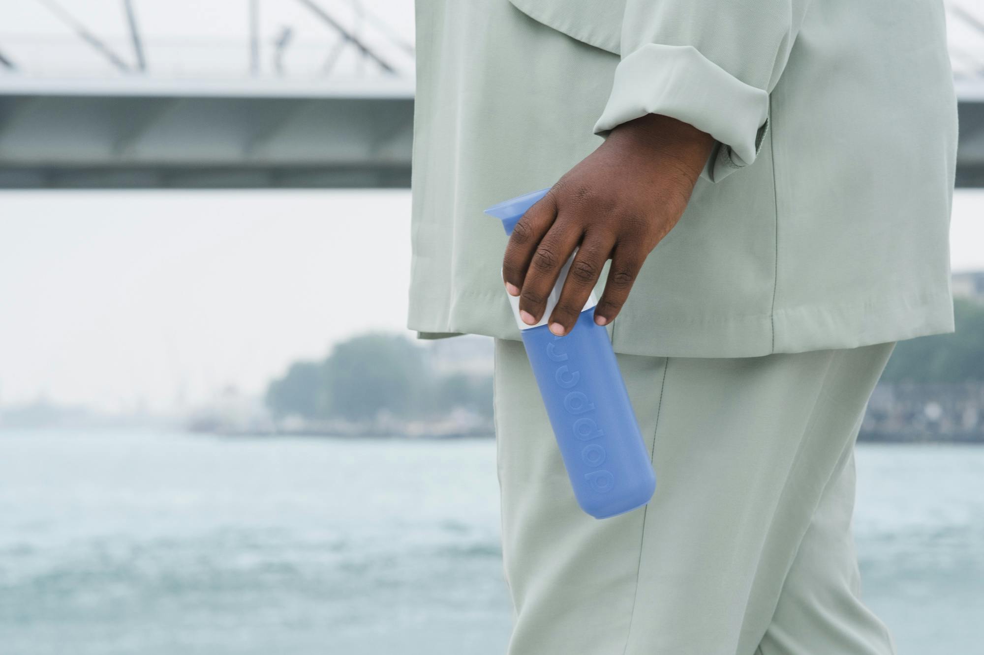 Man in light grey suit holding a blue Dopper bottle in front of a bridge and water